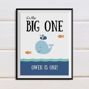 May include: A framed print with a whale illustration. The print features a light blue whale with a water spout, two orange fish, and the text "it's the BIG ONE" and "OWEN IS ONE!" against a white background. The frame is black.