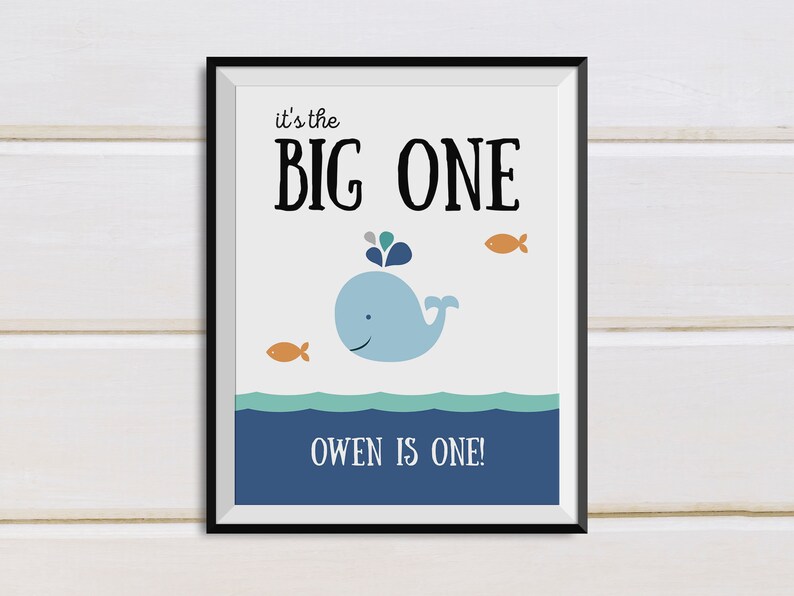 May include: A framed print with a whale illustration. The print features a light blue whale with a water spout, two orange fish, and the text "it's the BIG ONE" and "OWEN IS ONE!" against a white background. The frame is black.