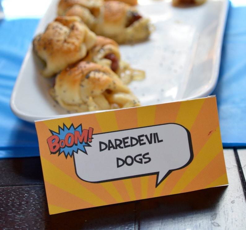 May include: A yellow and orange sign with a comic book style speech bubble that says "BOOM! DAREDEVIL DOGS". The sign is on a wooden table next to a white plate with several crescent-shaped pastries.