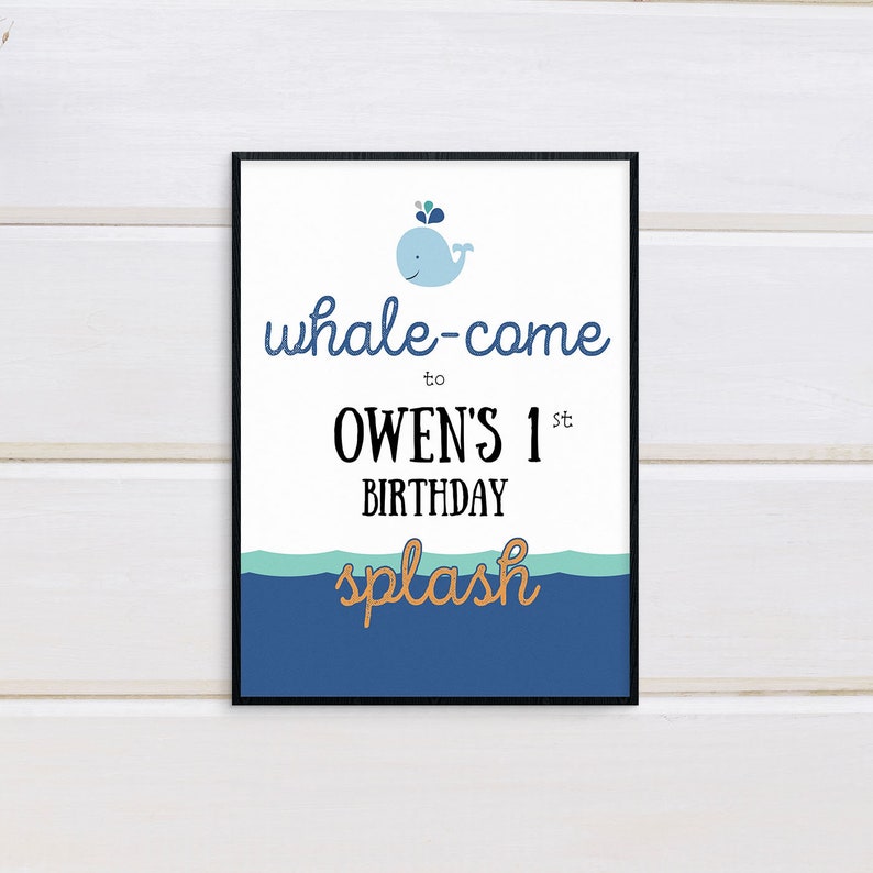 May include: A framed print with a white background and a blue whale illustration. The text reads "whale-come to Owen's 1st Birthday splash" in blue, black, and gold lettering. The bottom of the print features a blue and teal wave design.