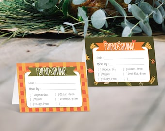 Puede incluir: Dos etiquetas de comida de Acción de Gracias de color naranja y marrón con el texto "FRIENDSGIVING!" y espacios en blanco para escribir el nombre del plato, quién lo hizo y las restricciones dietéticas. Las etiquetas tienen un patrón a cuadros y hojas de otoño.