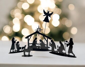 May include: Black metal nativity scene featuring silhouettes of the holy family, angels, and wise men. The figures are arranged under a star and a stable, creating a festive holiday decoration. The scene is set against a blurred background.