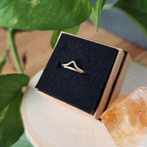 May include: A gold-colored ring with a simple, curved design, displayed in a small, brown box with black lining.