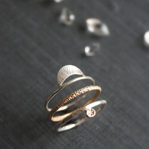 May include: A collection of silver and gold rings. One ring features a crescent moon design, while another has a textured band. A third ring has a small, round gold accent. The rings are displayed on a dark gray surface.
