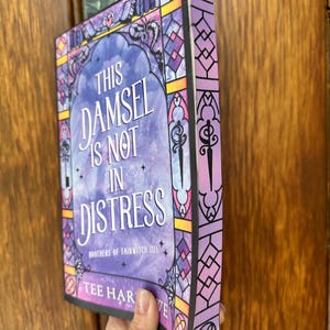 Op de afbeelding: Een boek getiteld "This Damsel Is Not In Distress" met een cover in glas-in-loodstijl. De cover heeft een paars en blauw kleurenschema met decoratieve elementen en de naam van de auteur. Het boek wordt voor een houten deur gehouden.