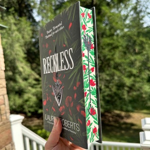 Puede incluir: Un libro de tapa dura titulado "Reckless" con una cubierta oscura con un emblema plateado y detalles florales rojos. El lomo tiene un patrón floral con flores rojas y hojas verdes. El libro se sostiene contra un fondo verde borroso.
