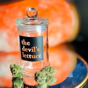 May include: A clear glass jar with a black label that reads "the devil's lettuce". The jar is sitting on a table with a piece of green leafy material in front of it.