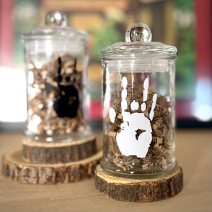 May include: Two glass jars with lids, one with a black handprint and the other with a white handprint. Both jars are filled with brown paper shreds and are sitting on wooden bases.