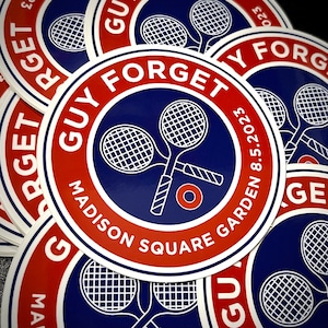 May include: Close-up of a stack of round stickers with a blue and red design. The stickers feature a tennis racket and the text "GUY FORGET MADISON SQUARE GARDEN 8.5.2023".