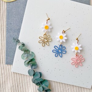 Könnte beinhalten: Drei Paar Hängeohrringe mit weißen Gänseblümchen-Anhängern und bunten Drahtblumen-Akzenten. Die Ohrringe werden auf einer weißen Oberfläche mit einem grünen Blattzweig präsentiert.