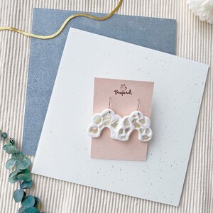 May include: White cloud-shaped earrings with a mosaic design, displayed on a pink card. The earrings have a unique, abstract shape and are set against a backdrop of textured paper and fabric.