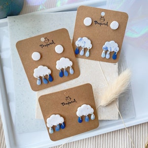 Peut inclure: Trois paires de boucles d'oreilles en argile polymère faites à la main, avec un nuage blanc et des gouttes de pluie bleues, et un cercle blanc avec le texte "teapeach".