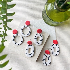 May include: A set of five pairs of handmade earrings. The earrings are made of white clay with black geometric patterns and red clay accents.