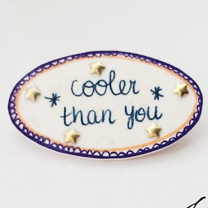 May include: A white oval pin with a purple border and gold stars. The pin says "cooler than you" in blue cursive writing.