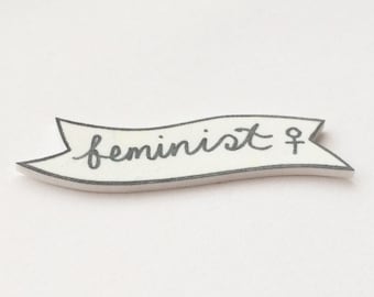May include: White banner with black outline and the word "feminist" written in cursive with a female symbol at the end.