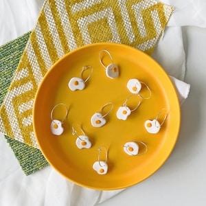 Könnte beinhalten: Ein gelber Teller mit verschiedenen weißen und gelben eiförmigen Ohrringen. Die Ohrringe sind aus Polymer Clay gefertigt und haben einen silbernen Ring.