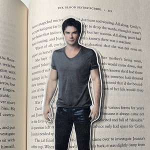 May include: A black and white bookmark featuring a photo of a man with dark hair and a black shirt. The bookmark is cut out in the shape of the man's body and is placed on a page of a book. The text on the page reads "INK BLOOD SISTER SCRIBE".