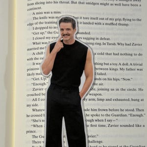 May include: A die-cut sticker of a man in a black sleeveless shirt and black pants, with his finger touching his face. The sticker is placed on top of a book with printed text.