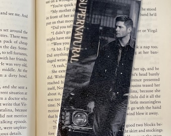 May include: A black and white photo of a man standing next to a car. The man is wearing a black jacket and has short brown hair. The car is a classic car with a large, round headlight. The photo is a bookmark with the word "SUPERNATURAL" printed on it.