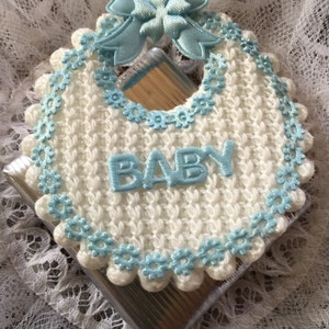May include: A white crocheted baby bib with a blue crocheted trim and a blue satin bow. The bib has the word "BABY" embroidered on it.