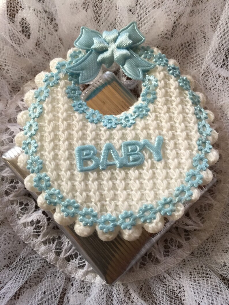 May include: A white crocheted baby bib with a blue crocheted trim and a blue satin bow. The bib has the word "BABY" embroidered on it.