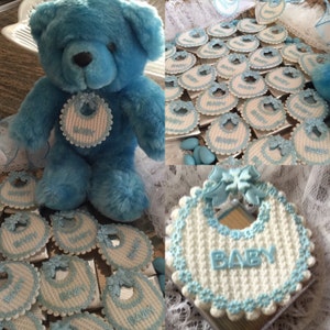 May include: Blue teddy bear wearing a white and blue crocheted bib.  The bear is surrounded by white and blue crocheted bibs with the word "BABY" on them.  The bibs are arranged on a silver tray.
