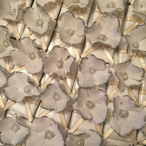 May include: A collection of white favour boxes with silver glitter trim and white paper flowers. Each box has a white satin ribbon tied around it.