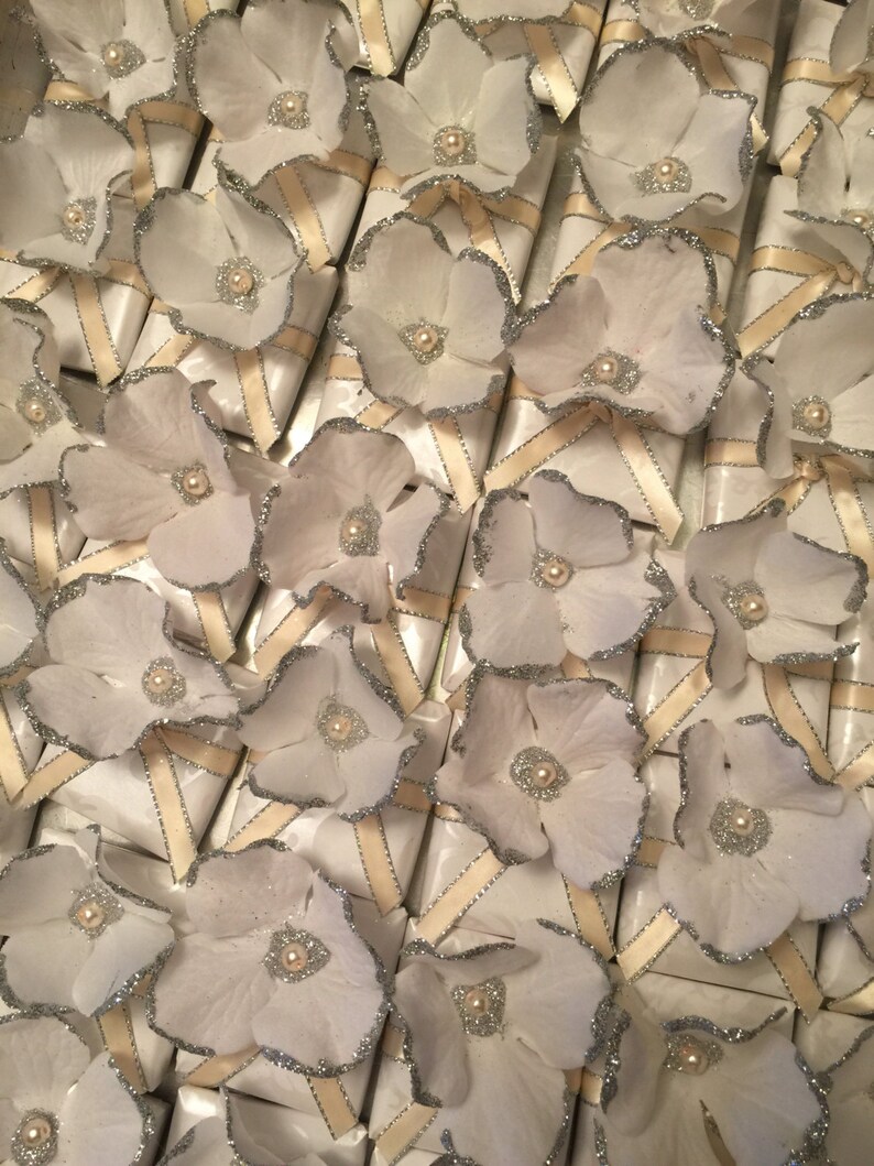 May include: A collection of white favour boxes with silver glitter trim and white paper flowers. Each box has a white satin ribbon tied around it.