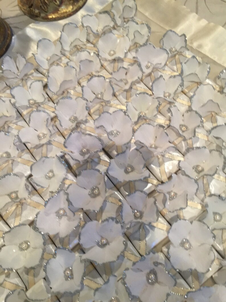 May include: A collection of white favour boxes decorated with white fabric flowers and silver glitter trim. The boxes are arranged in rows and are ready for a special occasion.