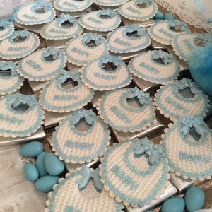 May include: A collection of blue and white baby bibs with the word "BABY" written on each one. The bibs are arranged in a grid pattern on a silver tray. The bibs are decorated with blue and white lace.