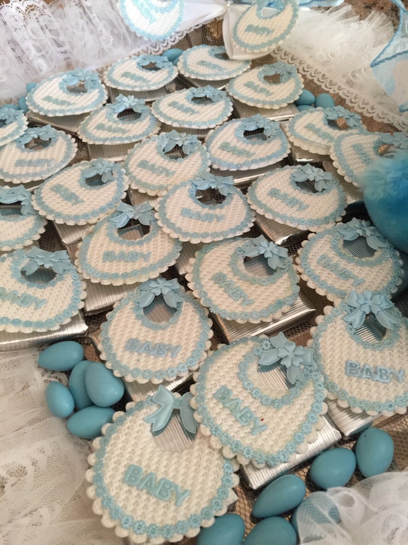 May include: A collection of blue and white baby bibs with the word "BABY" written on each one. The bibs are arranged in a grid pattern on a silver tray. The bibs are decorated with blue and white lace.