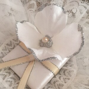 May include: A white gift box with a satin ribbon and a white flower with a pearl in the centre. The flower has silver glitter around the edges.