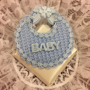 May include: A blue crocheted baby bib with a white satin bow and the word "BABY" in silver lettering. The bib is on top of a white satin box.