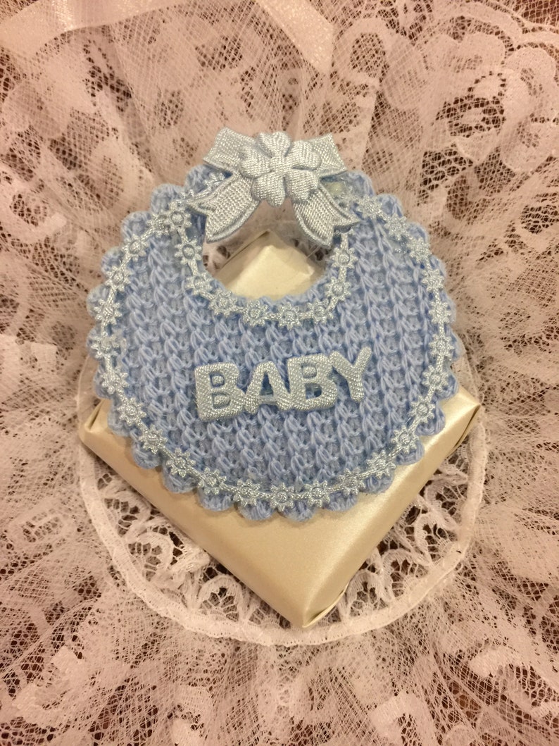 May include: A blue crocheted baby bib with a white satin bow and the word "BABY" in silver lettering. The bib is on top of a white satin box.