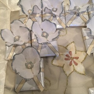 May include: White gift boxes with silver trim and a white flower on top. Each flower has a pearl in the centre. The boxes are tied with a cream ribbon.