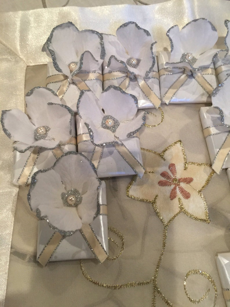 May include: White gift boxes with silver trim and a white flower on top. Each flower has a pearl in the centre. The boxes are tied with a cream ribbon.