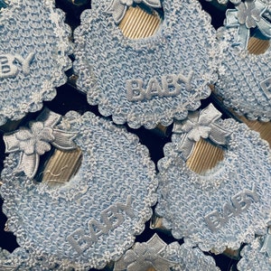 May include: A close-up of several blue crocheted baby bibs with a white lace trim and the word "BABY" embroidered on each bib.