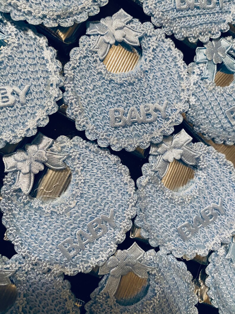May include: A close-up of several blue crocheted baby bibs with a white lace trim and the word "BABY" embroidered on each bib.