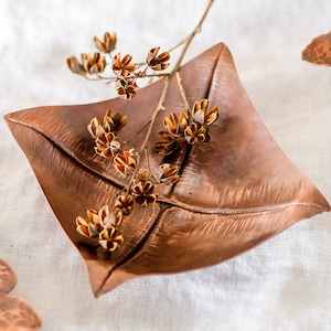 May include: A copper leaf-shaped dish with a hammered texture, featuring a sprig of dried flowers with brown seed pods.