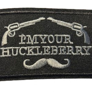 May include: Black rectangular patch with embroidered text and imagery. The patch features two silver revolvers, the phrase "I'M YOUR HUCKLEBERRY" in silver, and a silver moustache. The patch has a black border.