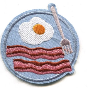 Può includere: Una toppa ricamata blu con un uovo fritto bianco, due fette di pancetta rosa e marrone e una forchetta argentata.