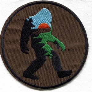 Op de afbeelding: Een bruin geborduurd patch met een zwart silhouet van Bigfoot die loopt. Het silhouet is gevuld met een groen, bruin en blauw landschap met een ondergaande zon.