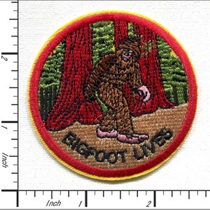 Op de afbeelding: Een ronde geborduurde patch met een rode rand en een gele buitenring. De patch toont een bruine Bigfoot die in een bos met rode en groene bomen loopt. De tekst "Bigfoot Lives" is in bruin op de onderkant van de patch geborduurd.