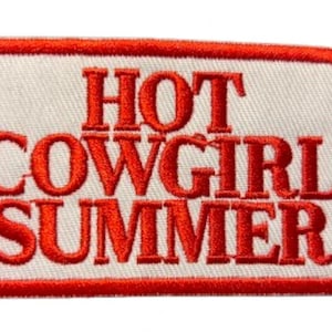 May include: Embroidered patch featuring the phrase "HOT COWGIRL SUMMER" in red, capitalised letters. The text is on a white rectangular background with a red border. The patch is designed to be sewn or ironed onto clothing or accessories.