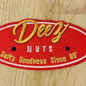 May include: Red oval embroidered patch with white and gold stitching. The patch says "Deez NUTS" in a stylized font and "Salty Goodness Since 69'" below.