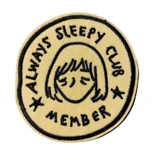 May include: A beige embroidered patch with black stitching. The patch is round and features a cartoon illustration of a person with their eyes closed. The text around the illustration reads "Always Sleepy Club Member".