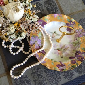 May include: A white porcelain serving dish with a gold rim and floral design. The dish features two hummingbirds and a gold stand. A string of white pearls is draped over the dish.