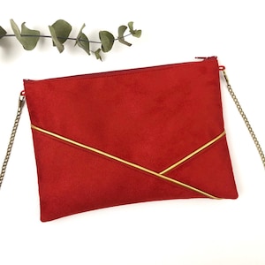 May include: A red suede clutch bag with a gold chain strap and gold accents. The bag has a geometric design with two gold lines forming a V shape.