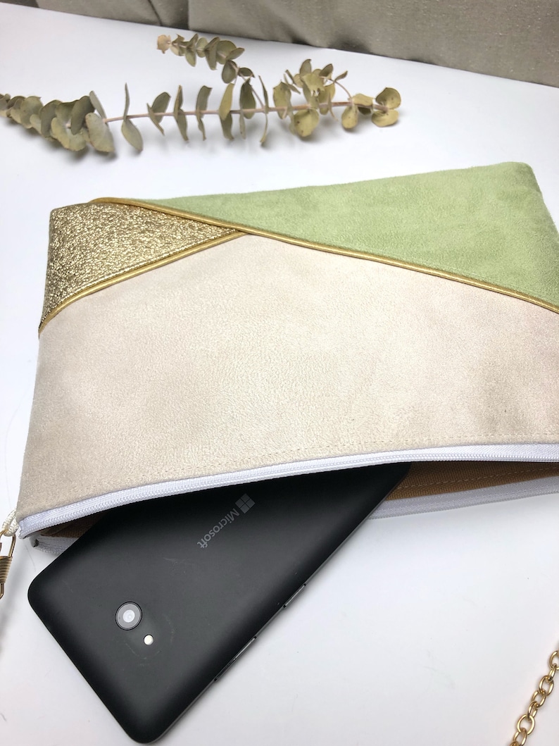 K&ouml;nnte beinhalten: Eine kleine Clutch in Hellgr&uuml;n, Beige und Gold mit Rei&szlig;verschluss und einer goldenen Kettenkette. Ein schwarzes Smartphone mit dem Microsoft-Logo befindet sich in der Clutch.