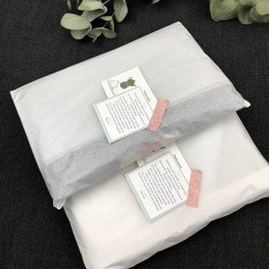 K&ouml;nnte beinhalten: Zwei wei&szlig;e Seidenpapierpakete mit einem rosa-wei&szlig;en Etikett mit der Aufschrift "These LAE" und einem blau-wei&szlig;en Etikett mit der Aufschrift "Thealouise".